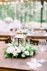 Wedding decorations in rustic style on wooden table, flower arrangement and golden lantern. Table served with cutlery and crockery, wineglasses.