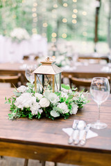 Wedding decorations in rustic style on wooden table, flower arrangement and golden lantern. Table served with cutlery and crockery, wineglasses.