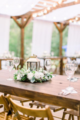 Wedding decorations in rustic style on wooden table, flower arrangement and golden lantern. Table served with cutlery and crockery, wineglasses.
