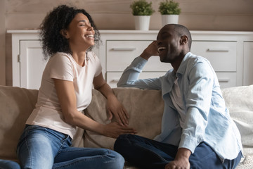 Overjoyed millennial african american married couple sitting on sofa, chatting.