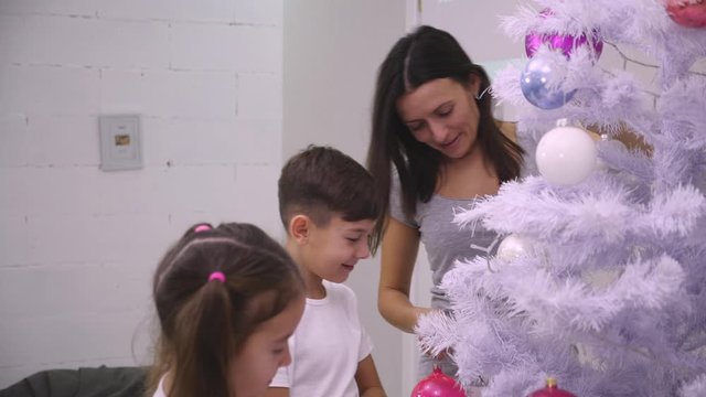 Mother and two kids are decorating a Christmas tree with colourful boubles in the living-room.