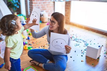 Young beautiful psicologist and toddler doing therapy using emoji emotions at kindergarten - obrazy, fototapety, plakaty