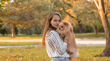 A young blonde smiles and holds a dog in her arms. Pomeranian spitz for a walk in the autumn park. Love to the animals