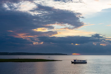 Fototapeta premium Wooden traditional boat sailing on Tapajos river at dusk with clouds and sunset in the background. Amazon, Brazil. Environment, climate change, travel, tourism and peace concept.