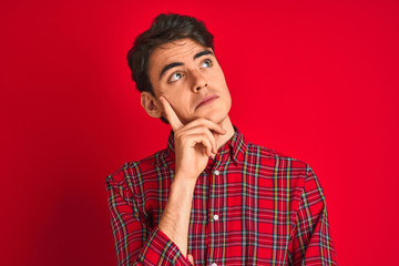 Teenager boy wearing red shirt standing over isolated background with hand on chin thinking about question, pensive expression. Smiling with thoughtful face. Doubt concept.