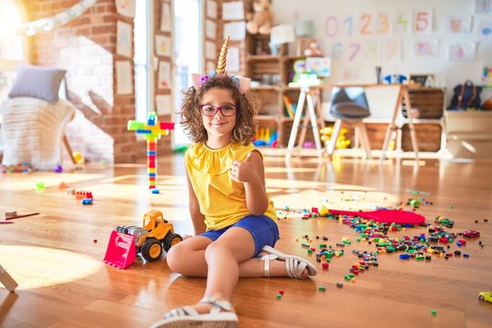 Beautiful toddler wearing glasses and unicorn diadem sitting on the floor at kindergarten doing happy thumbs up gesture with hand. Approving expression looking at the camera with showing success.