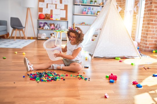 Beautiful toddler wearing glasses and unicorn diadem sitting on the floor playing with building blocks at kindergarten