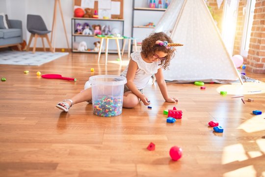 Beautiful toddler wearing glasses and unicorn diadem sitting on the floor playing with building blocks at kindergarten