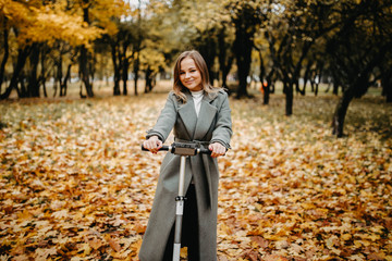Woman in a coat in autumn on an electric scooter in an autumn park. Riding on electric vehicles in cold weather.