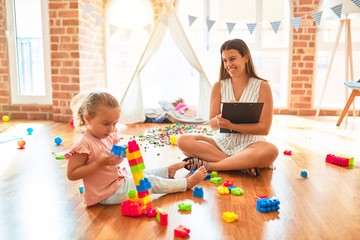 Fototapeta premium Beautiful psycologist and blond toddler girl doing therapy building tower using plastic blocks at kindergarten