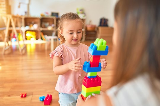 Beautiful teacher and blond toddler girl building tower using plastic blocks at kindergarten