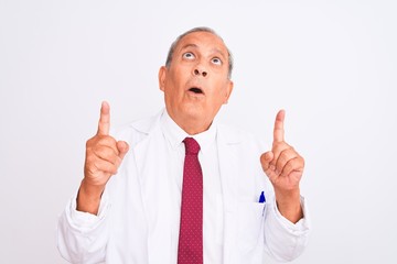 Senior grey-haired scientist man wearing coat standing over isolated white background amazed and surprised looking up and pointing with fingers and raised arms.