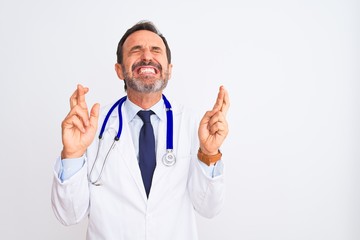 Middle age doctor man wearing coat and stethoscope standing over isolated white background gesturing finger crossed smiling with hope and eyes closed. Luck and superstitious concept.