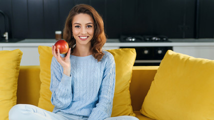 Beautiful young woman on diet holds fresh red apple and having rest in the stylish modern room.