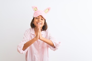 Beautiful child girl wearing sleep mask and pajama standing over isolated white background begging...