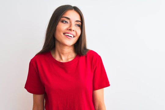 Young beautiful woman wearing red casual t-shirt standing over isolated white background looking away to side with smile on face, natural expression. Laughing confident.