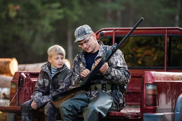  Vader en zoon zitten in een pick-up na de jacht in het bos. Papa die jongensmechanisme van een jachtgeweergeweer toont. © romankosolapov