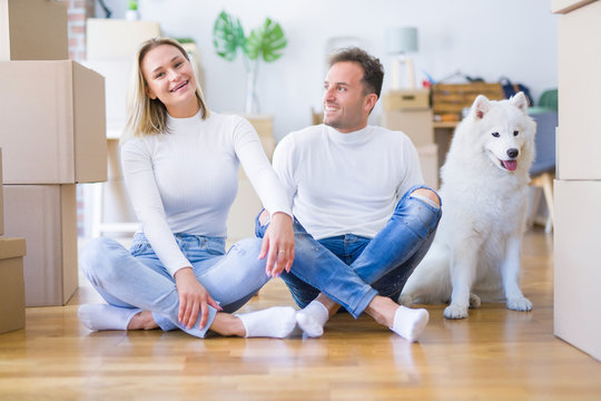Young beautiful couple with dog sitting on the floor at new home around cardboard boxes