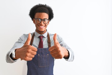 Young african american shopkeeper man wearing apron glasses over isolated white background approving doing positive gesture with hand, thumbs up smiling and happy for success. Winner gesture.