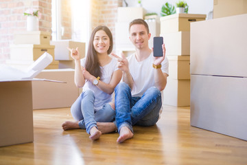 Beautiful couple sitting on the floor holding smartphone at new home around cardboard boxes very happy pointing with hand and finger to the side