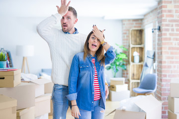 Young beautiful couple standing at new home around cardboard boxes surprised with hand on head for...