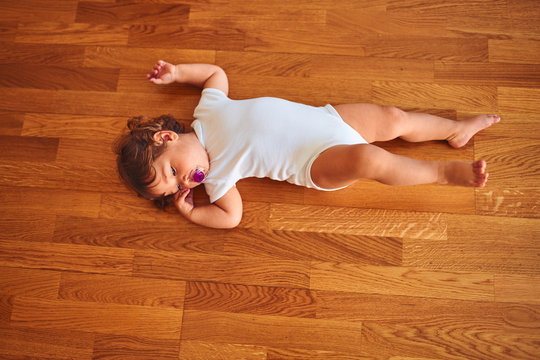 Beautiful toddler child girl wearing white bodysuit lying down on the floor using pacifier