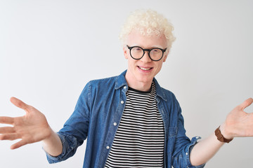 Young albino blond man wearing denim shirt and glasses over isolated white background smiling cheerful offering hands giving assistance and acceptance.