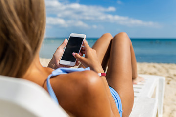 Closeup of young girl lying on beach with mobile phone in hand on tropical island. Girl on vacation at sea uses a smartphone and a social network on a background of sea waves