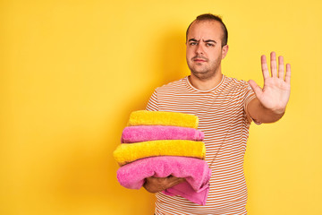 Young shopkeeper man holding folded towels standing over isolated yellow background with open hand doing stop sign with serious and confident expression, defense gesture