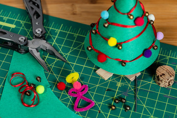 A tightly framed shot of a construction paper Christmas tree decorated with pom-poms, ribbon and bells.  Scattered on the cutting mat are various other supplies and tools..