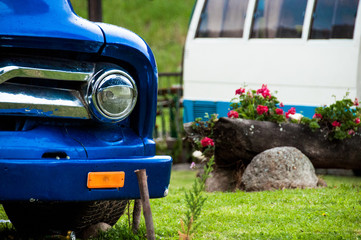 faro de carro azul estacionado en un jardín con mas plantas y un camión al fondo