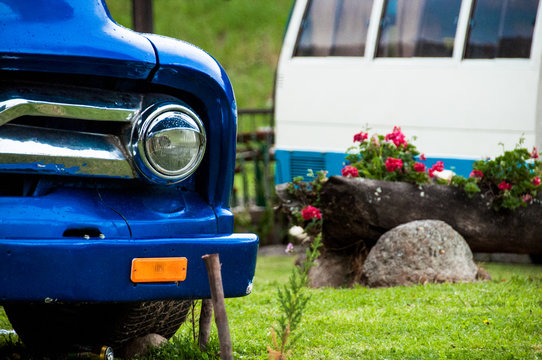 faro de carro azul estacionado en un jard&iacute;n con mas plantas y un cami&oacute;n al fondo