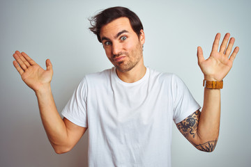 Fototapeta premium Young handsome man with tattoo wearing casual t-shirt over isolated white background clueless and confused expression with arms and hands raised. Doubt concept.