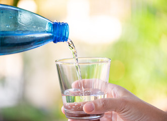 Hand holding blue water bottle pouring water into the glass on blurry natural green tree background.Health care food and drink concept.