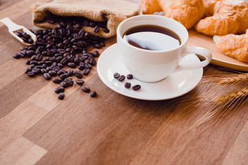 Top view white hot coffee cup and beans on wooden table with croissant bread background.Black coffee menu for breakfast in the coffee shop.Selective focus.