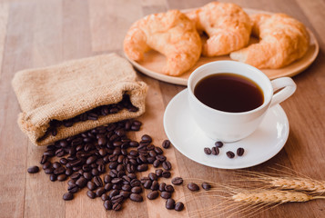 Top view white hot coffee cup and beans on wooden table with croissant bread background.Black coffee menu for breakfast in the coffee shop.Selective focus.