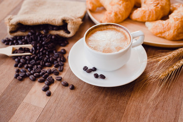 Top view white hot coffee cup and beans on wooden table with croissant bread background.Latte art coffee menu for breakfast in the coffee shop.Selective focus.