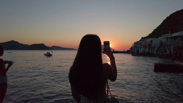 4K Silhouette of a girl taking photos with mobile at sunset from Klima, Milos, Greece.