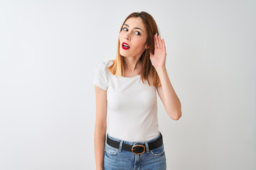 Beautiful redhead woman wearing casual t-shirt standing over isolated white background smiling with hand over ear listening an hearing to rumor or gossip. Deafness concept.