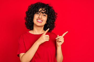 Young arab woman with curly hair wearing casual t-shirt over isolated red background smiling and looking at the camera pointing with two hands and fingers to the side.