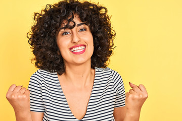 Young arab woman with curly hair wearing striped dress over isolated yellow background screaming proud and celebrating victory and success very excited, cheering emotion