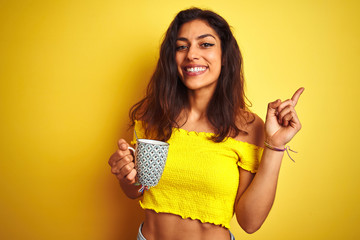 Young beautiful woman drinking cup of coffee standing over isolated yellow background very happy pointing with hand and finger to the side