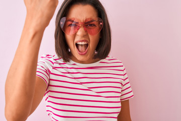 Young beautiful woman wearing fashion sunglasses with hearts over isolated pink background annoyed and frustrated shouting with anger, crazy and yelling with raised hand, anger concept