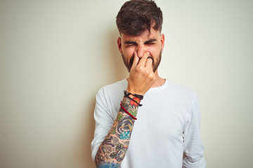 Young man with tattoo wearing t-shirt standing over isolated white background smelling something...