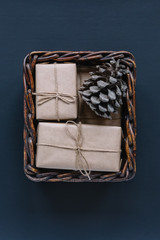 Vertical close up photo of wooden basket with christmas gifts in kraft paper and fir cone. Blue background. Home decor. Preparation for celebration. Flat lay. Happy New Year and Christmas concept. 