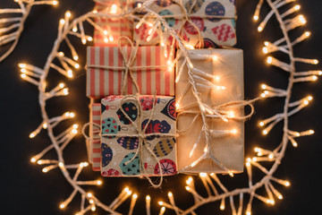 Fototapeta premium A bunch of Christmas gift boxes in multicolored kraft wrap under the garland with warm light. Selective focus on the top gift. Preparation for celebrating. Merry Christmas and Happy New Year concept.