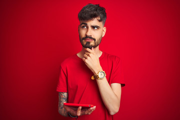 Young man with tattoo using tablet standing over isolated red background serious face thinking...