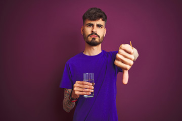 Young man with tattoo drinking glass of water standing over isolated purple background with angry...