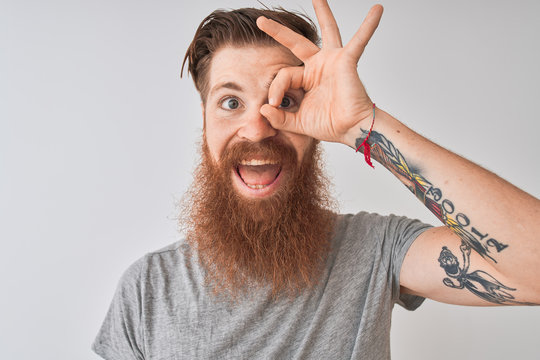 Young redhead irish man wearing t-shirt standing over isolated grey background with happy face smiling doing ok sign with hand on eye looking through fingers