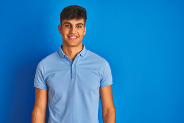 Young indian man wearing casual polo standing over isolated blue background with a happy and cool smile on face. Lucky person.
